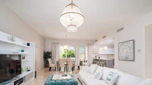 Un salon moderne avec un canapé blanc, un pouf bleu, un coin repas avec une table et des chaises, une cuisine en arrière-plan et un lustre.