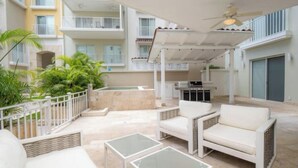 Une terrasse extérieure couverte, dotée de mobilier blanc, une piscine et une vue sur un immeuble à plusieurs étages.