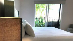 A neatly made bed with white linens and two pillows, a bedside lamp, and a view of a balcony with a railing and greenery outside. - Exotic Getaway 2BR in Plaza Puntarena (Rio Hato)
