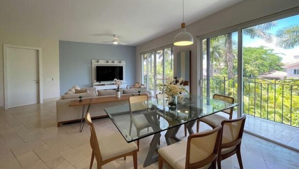 A spacious living area with a glass dining table, comfortable seating, a large TV, and a balcony with a view of greenery. - Exotic Getaway 2BR in Plaza Puntarena (Rio Hato)