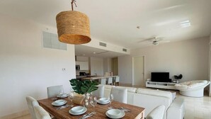Un espace repas moderne avec une table en bois dressée pour quatre personnes, entourée de chaises blanches. Un luminaire suspendu en fibres tressées est accroché au-dessus. Au fond, on aperçoit un salon avec un canapé blanc et un téléviseur à écran plat.