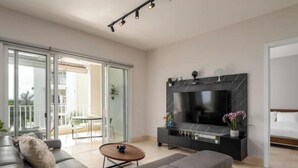 Una sala de estar moderna con un sofá gris, un televisor de pantalla plana sobre un mueble negro y un ventanal corredero que da a un área exterior.