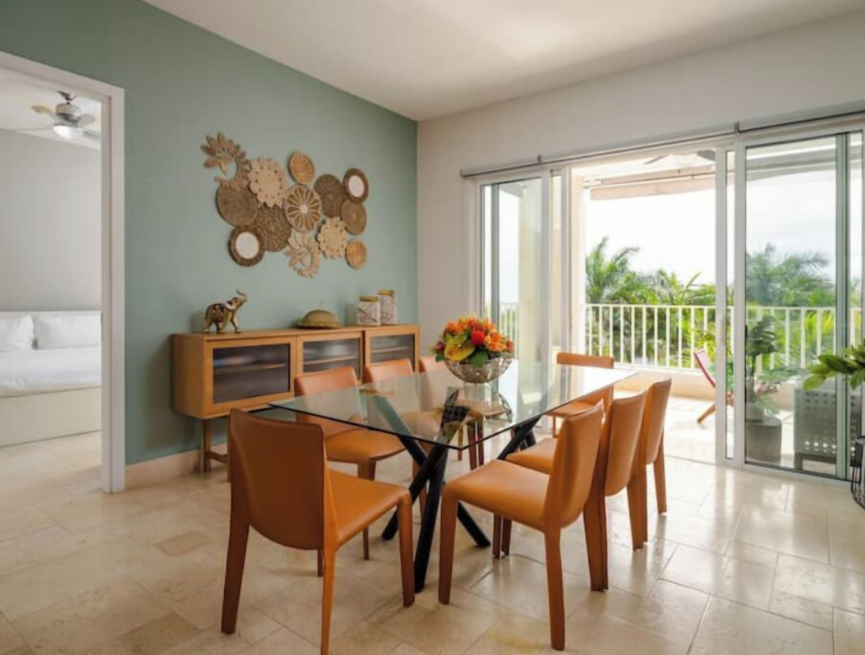 A dining area with a glass table and wooden chairs, a decorative wall, and a balcony with a view of greenery.