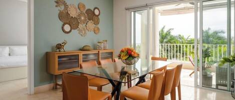 Un comedor con mesa de vidrio y sillas de madera, una pared decorativa y un balcón con vistas a la vegetación.