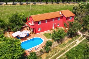 Une maison rouge avec une piscine, entourée de verdure et dotée d’une allée pavée.