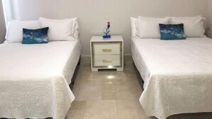 Ein Hotelzimmer mit zwei Betten, jeweils mit weißer Bettwäsche und blauen Kissen. An einem kleinen Nachttisch steht eine Lampe und ein dekorativer Gegenstand.