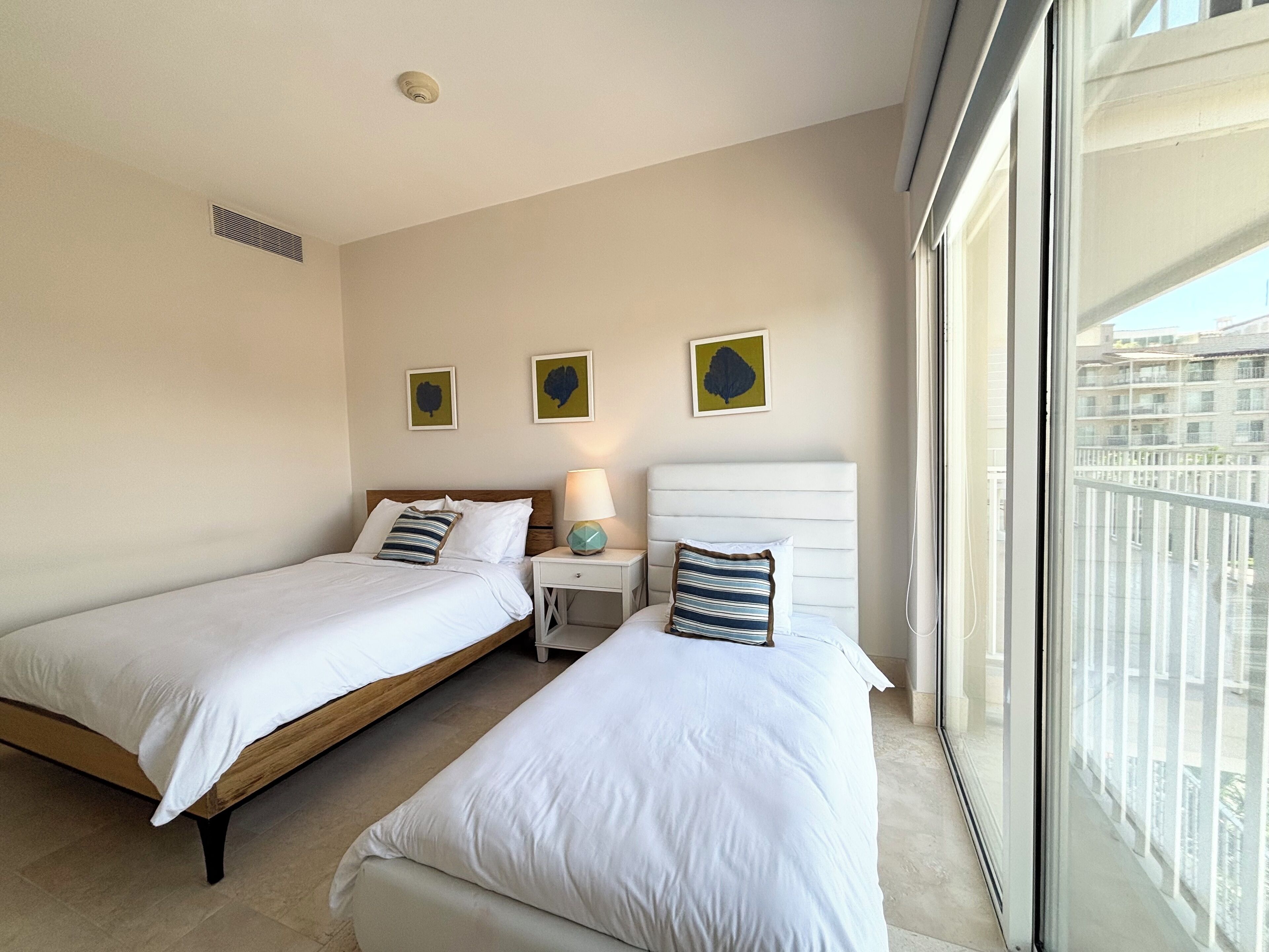 A hotel room with two beds, a nightstand, a lamp, and a window with a view of buildings.