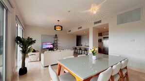 A modern living room with a dining area, a kitchen, and a TV in the background. - Tropical Retreat 2BR in Plaza Puntarena (Rio Hato)