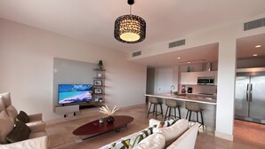 A modern living room with a sofa, a coffee table, and a TV. There is a kitchen area with a refrigerator, microwave, and bar stools. - Tropical Retreat 2BR in Plaza Puntarena (Rio Hato)