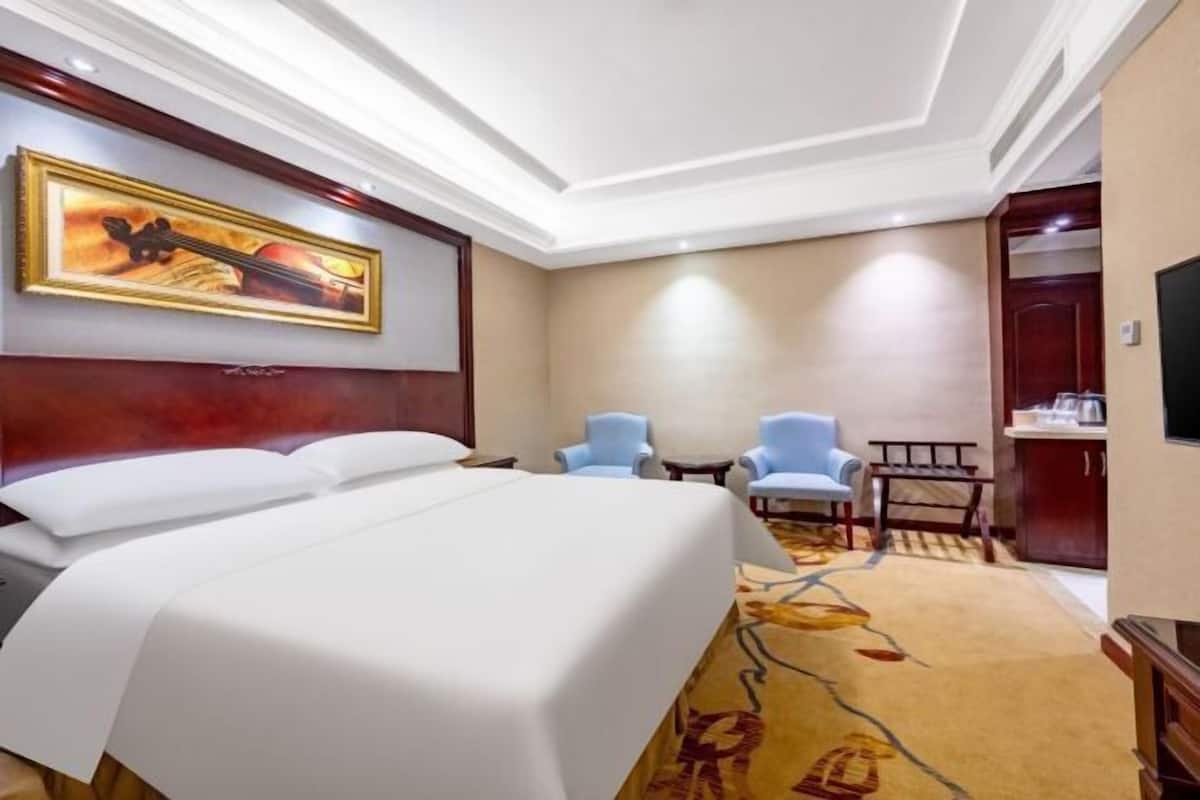 A hotel room with a large bed, two armchairs, a small table, a painting on the wall, and a television.