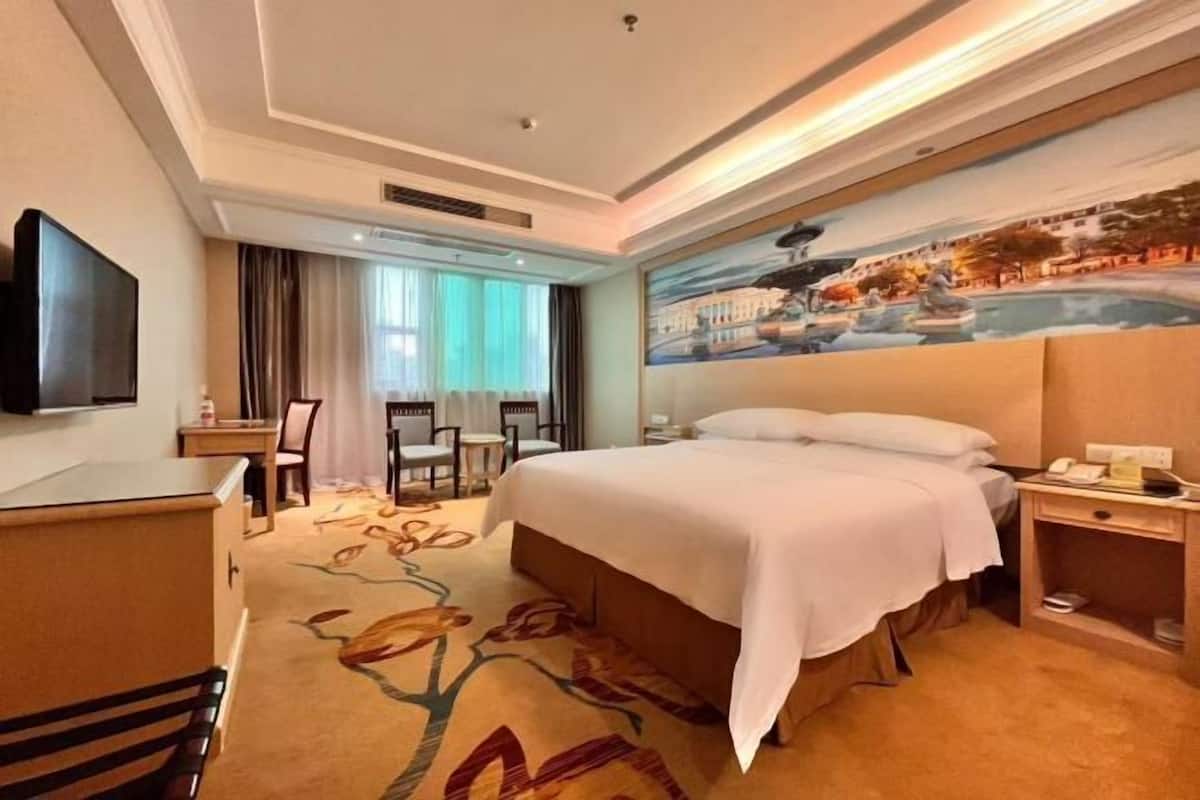 A hotel room with a large bed, a desk, a chair, a TV, and a painting of a landscape.