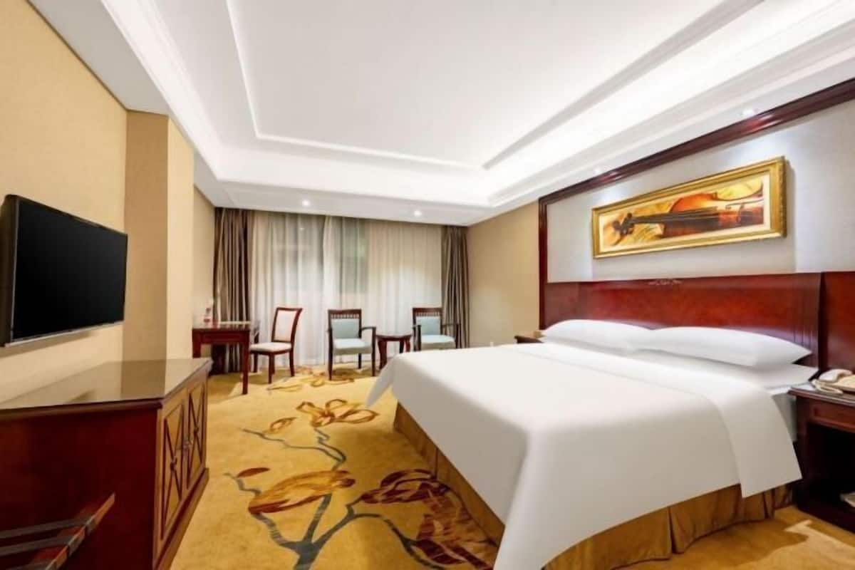 A hotel room with a large bed, a desk with a flat-screen TV, a painting on the wall, and a window with curtains.