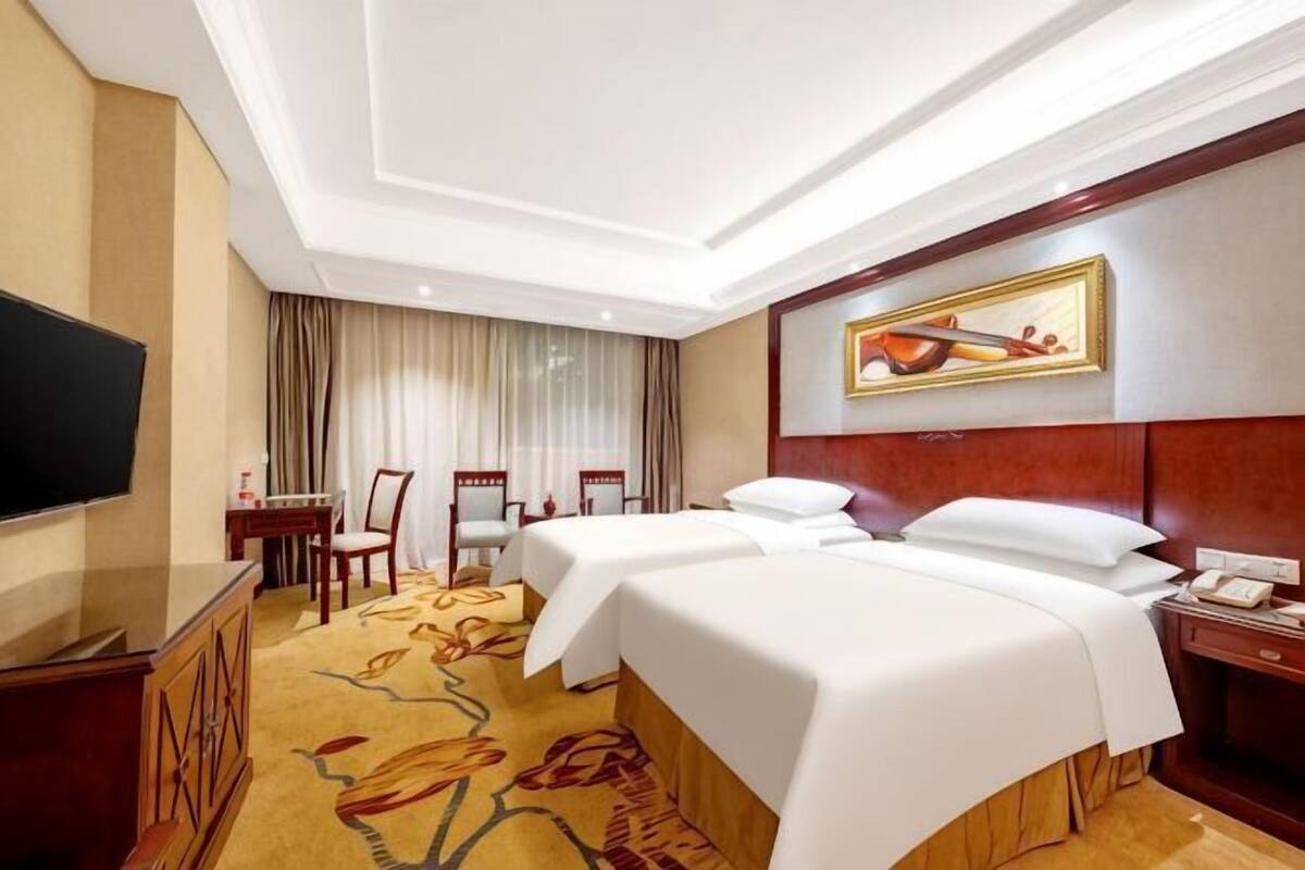 A hotel room with two beds, a television, a desk with a chair, and a painting on the wall.