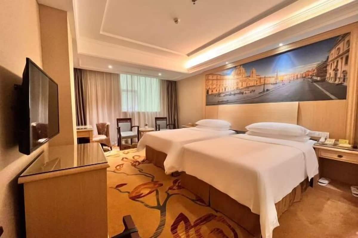 A hotel room with two beds, a TV, a desk, a chair, and a painting of a cityscape on the wall.