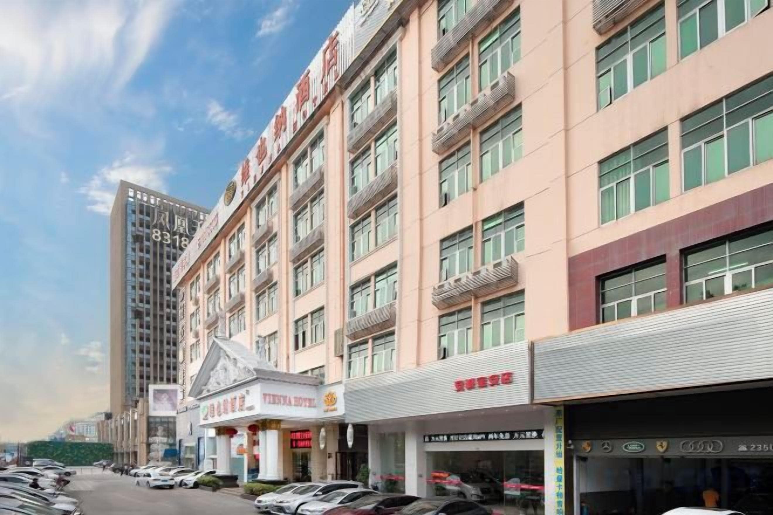 A multi-story hotel with a row of shops on the ground floor, a parking lot in front, and a tall building in the background.