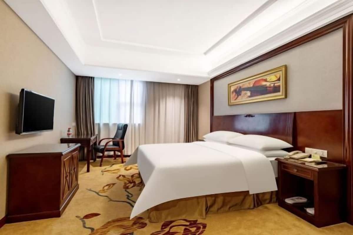 A hotel room with a bed, a desk, a chair, a television, and a painting on the wall.