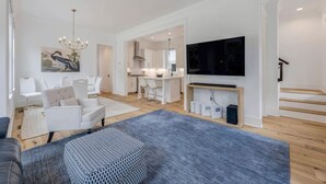Ett rymligt vardagsrum med en grå soffa, vita fåtöljer, en kristallkrona och en platt-TV som är monterad på väggen.