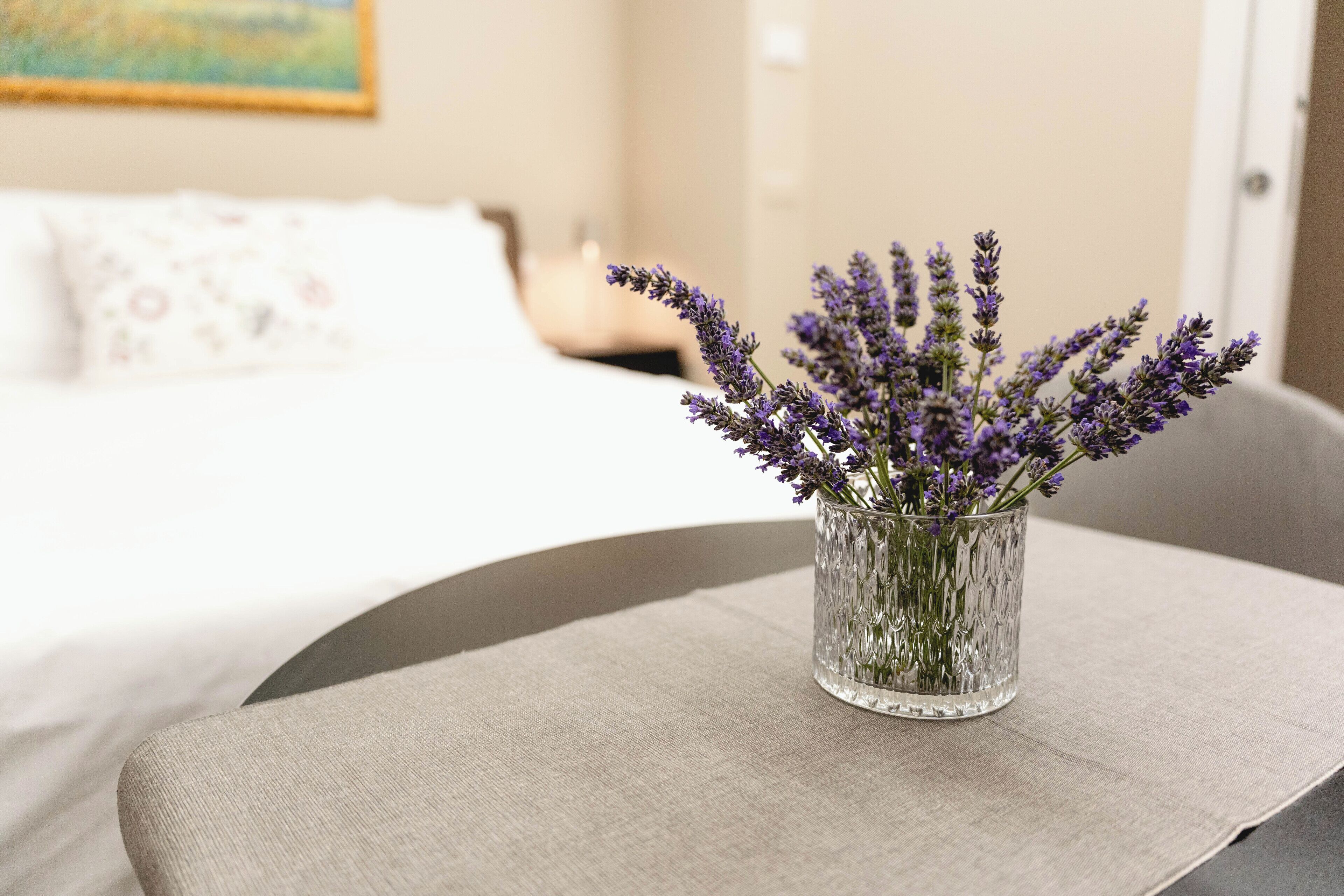 Una habitación con una cama, una mesita con un jarrón de flores de lavanda y un cuadro en la pared.