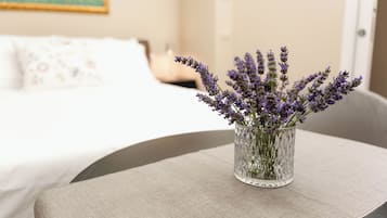 Une pièce avec un lit, une petite table sur laquelle est posé un vase de fleurs de lavande, et un tableau accroché au mur.