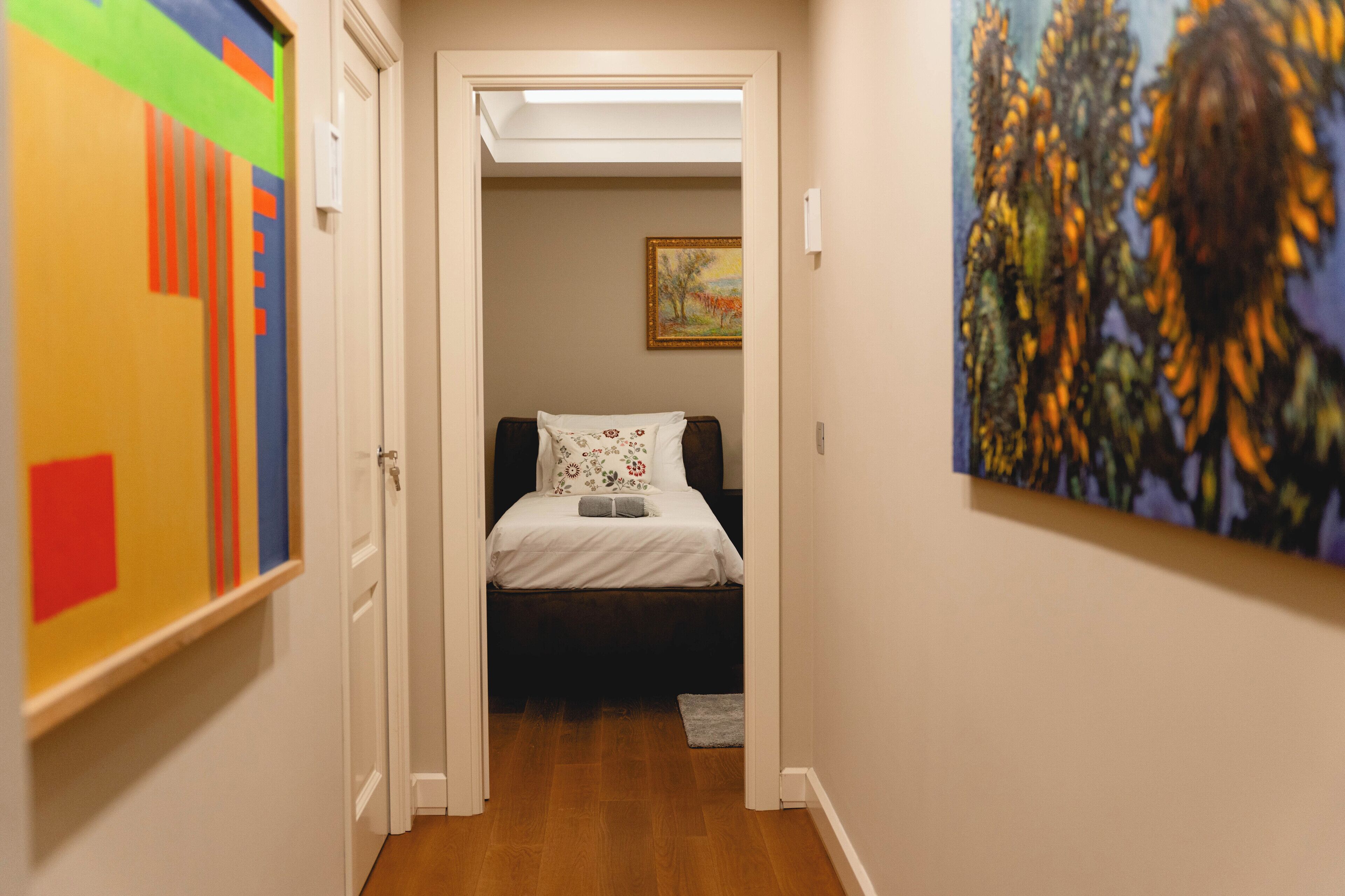 A narrow hallway leading to a bedroom with a bed, a painting on the wall, and a door on the left.