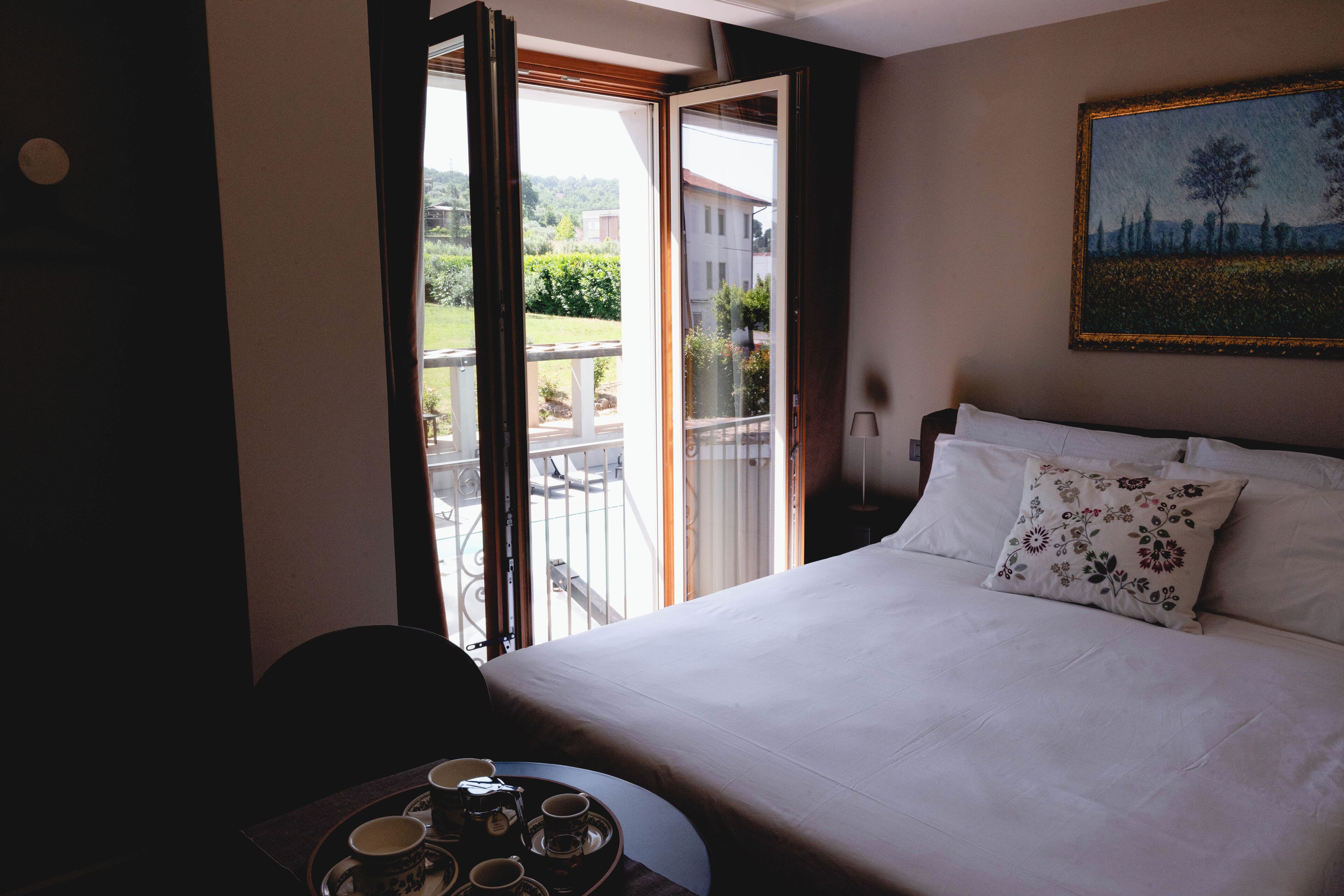 A hotel room with a bed, a table with cups and saucers, and a balcony with a view of the countryside.