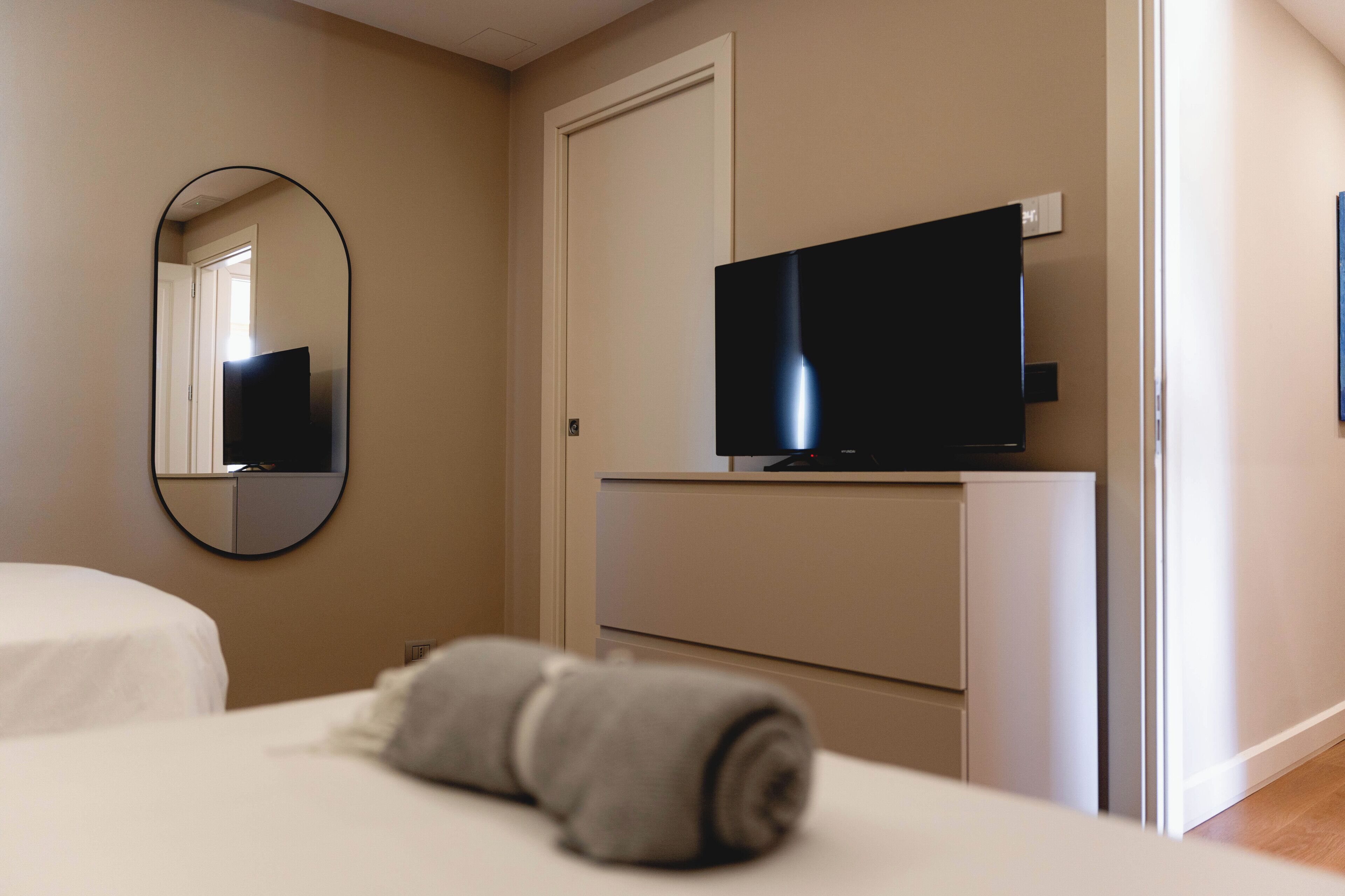 A hotel room with a bed, a TV mounted on a cabinet, a mirror on the wall, and a rolled towel on the bed.