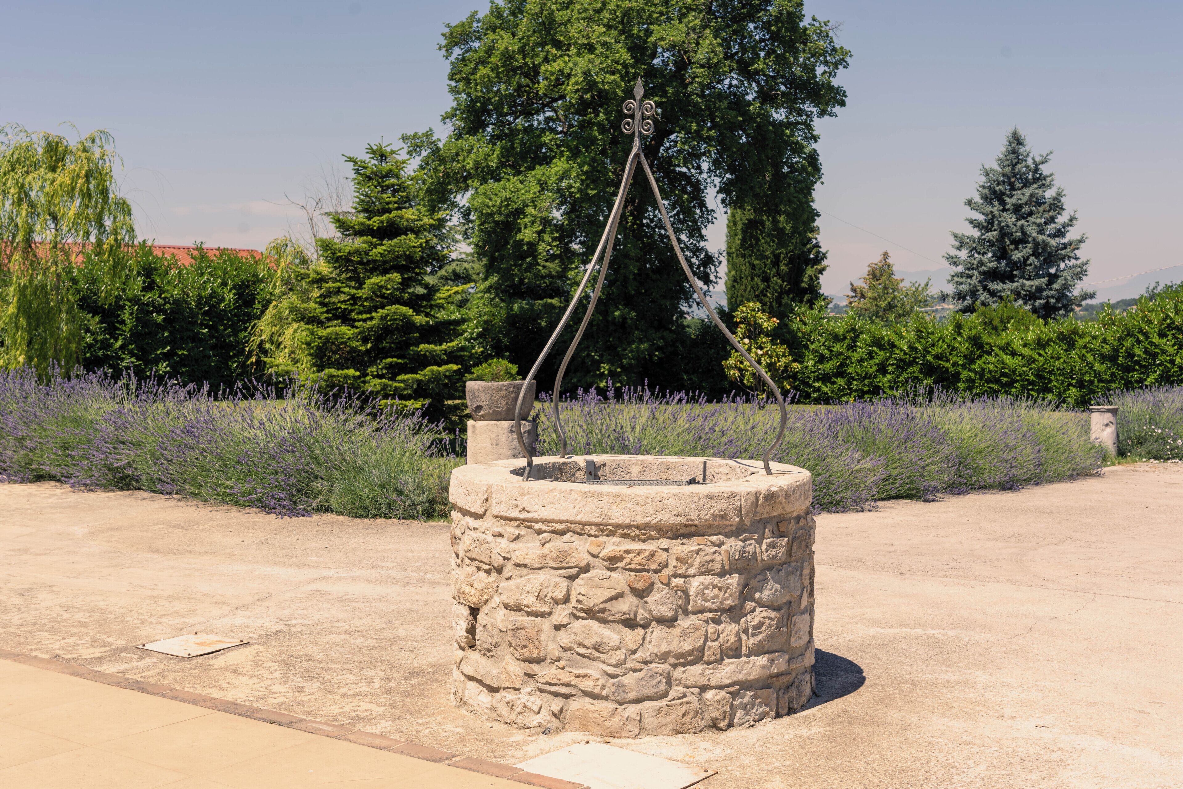 A stone well with a metal structure in a landscaped area with trees and lavender.