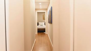 A narrow hallway with wooden flooring and white walls, leading to a bedroom with a bed and a painting on the wall.