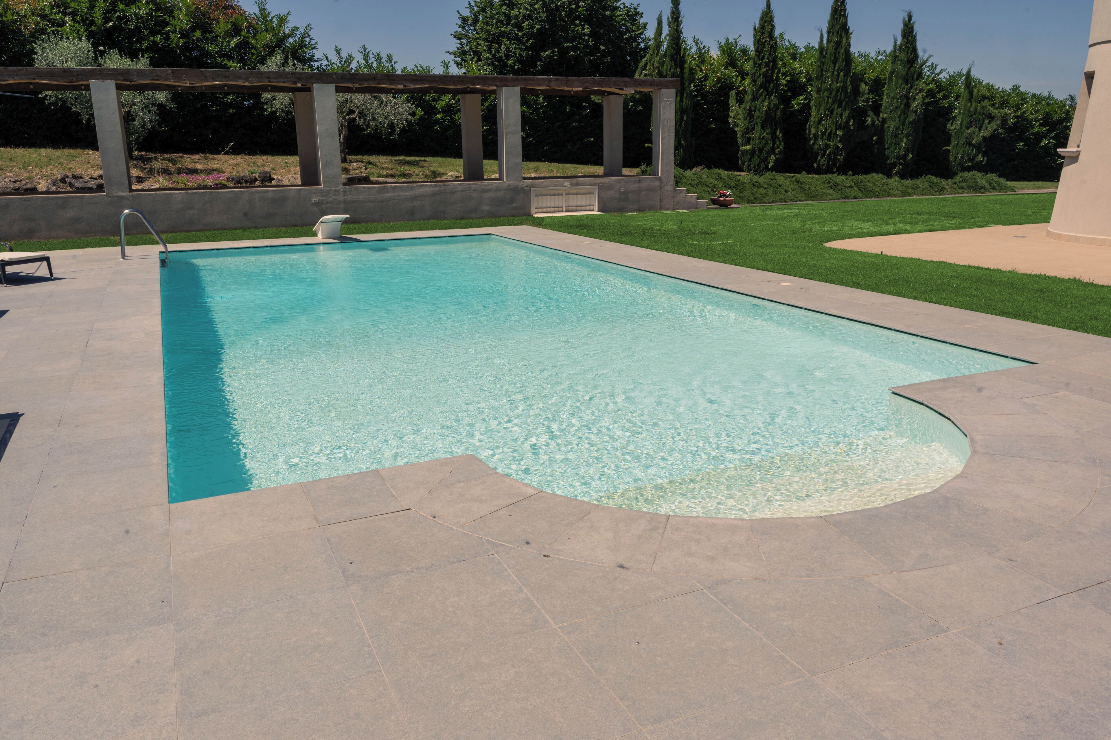 Piscina retangular com água azul cristalina, ladeada por um deck de concreto e uma pérgula de madeira.