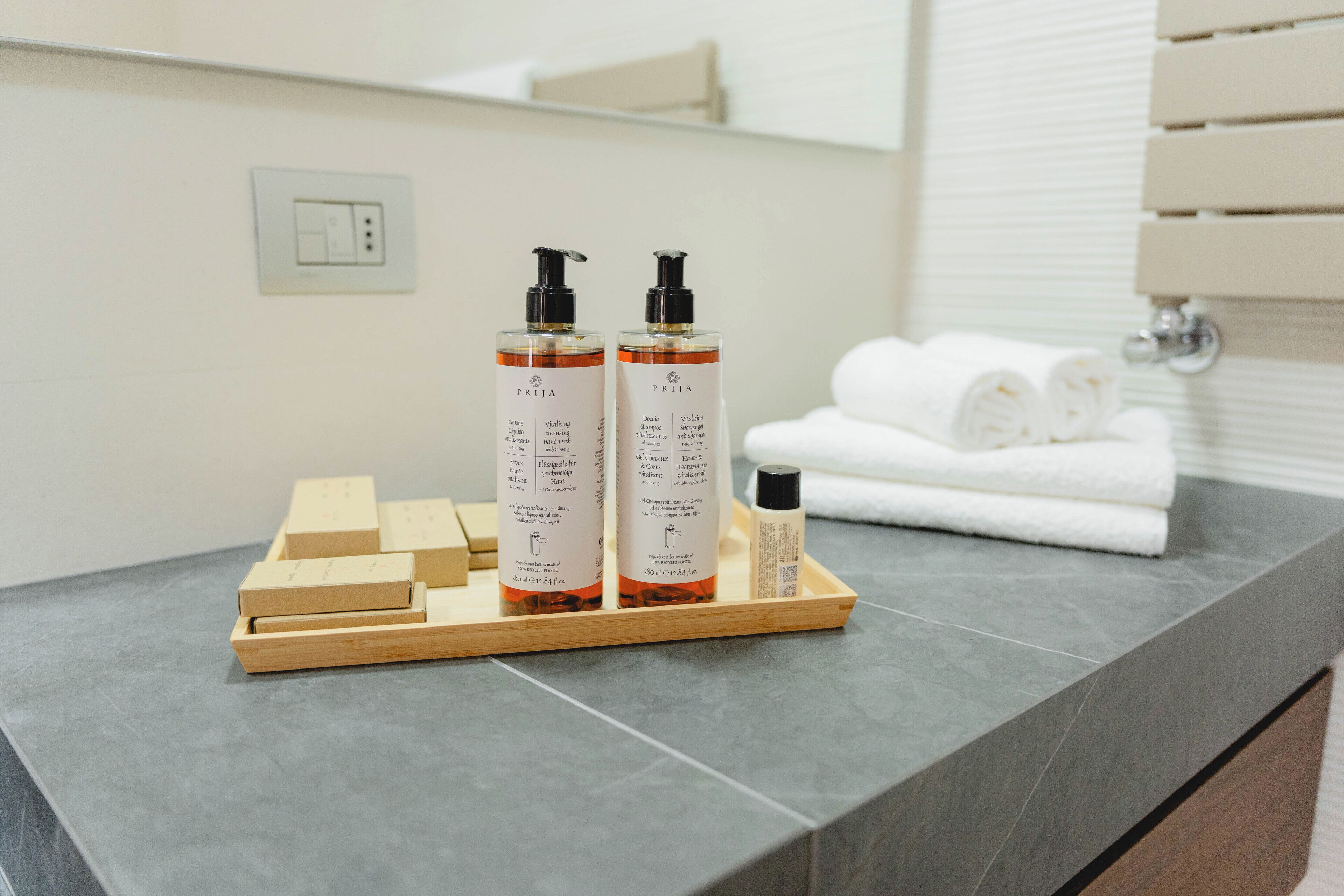 A bathroom with a grey countertop, two bottles of PRIJA body wash, a small bottle of lotion, and neatly folded white towels.