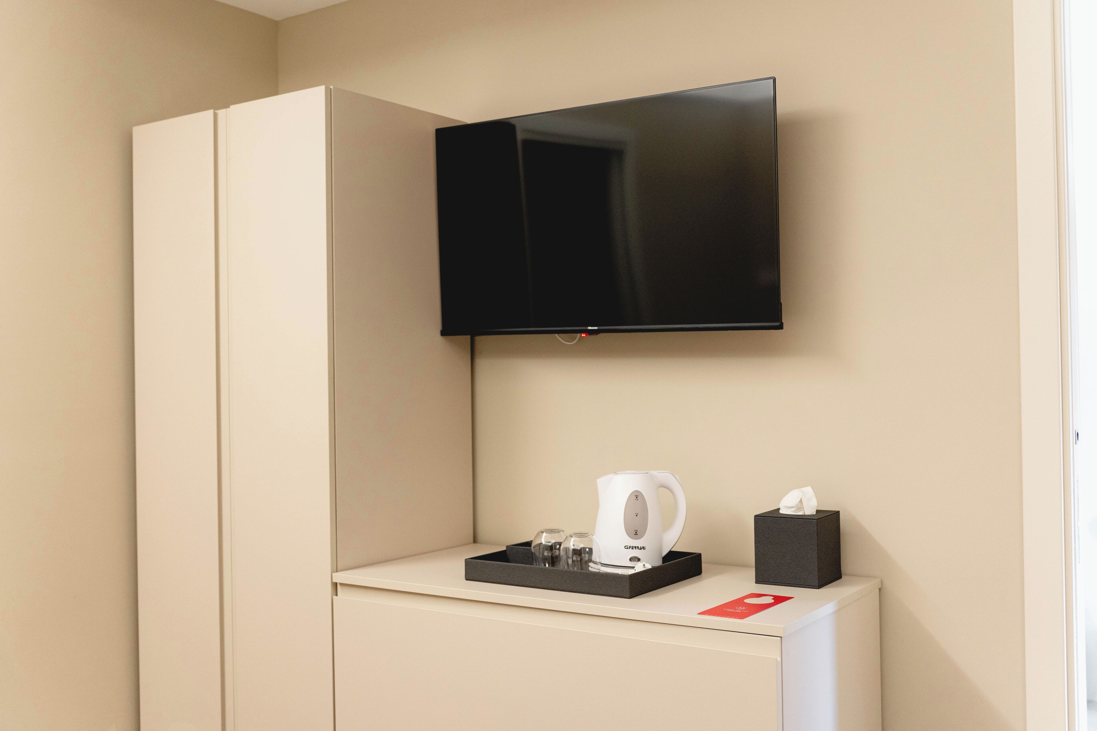 A hotel room with a flat-screen TV mounted on the wall, a minibar with a coffee maker, and a tissue box.