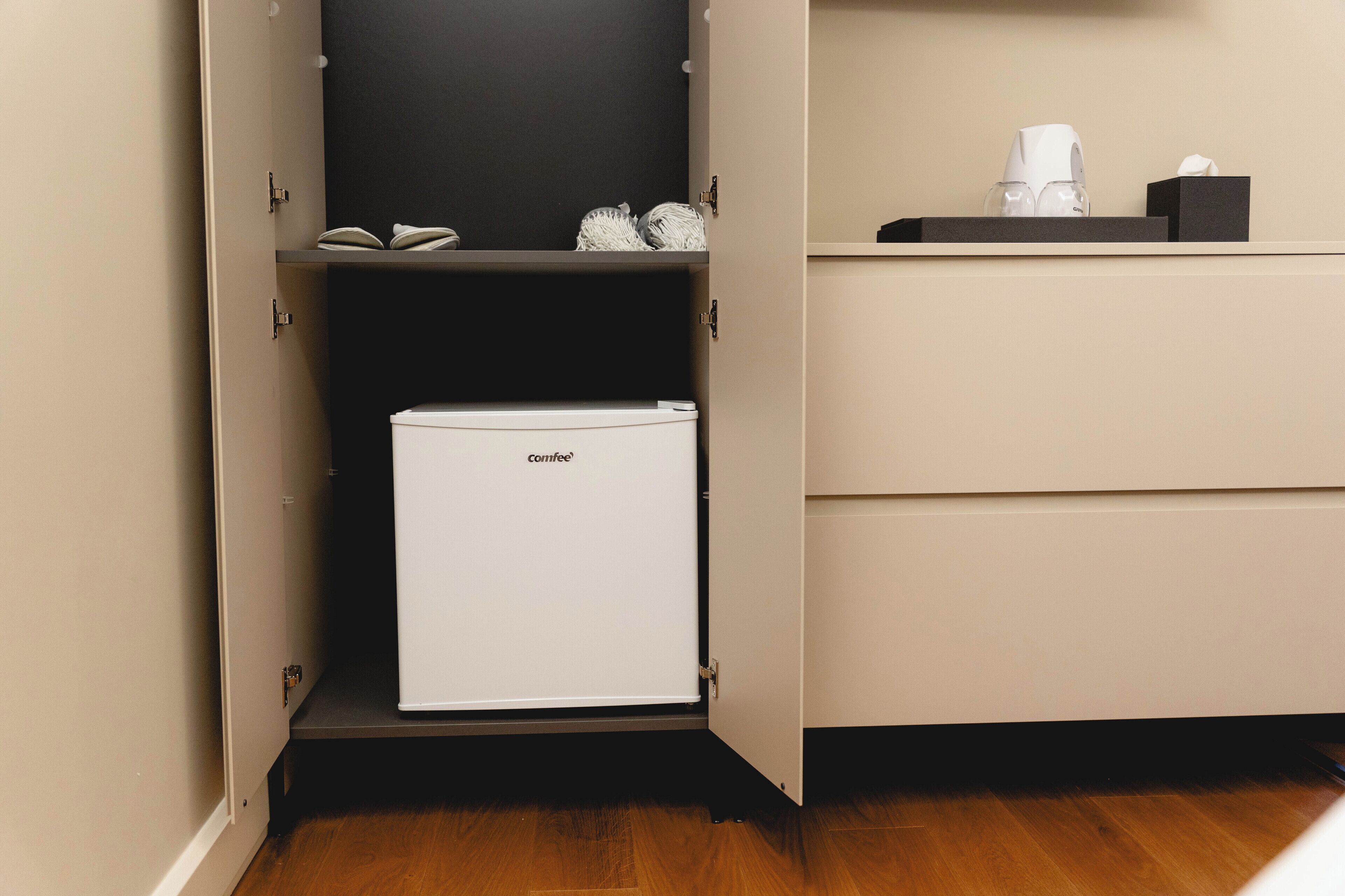 A compact kitchenette with a small refrigerator, a shelf with a book and a glass, and a countertop with a coffee maker and tissues.