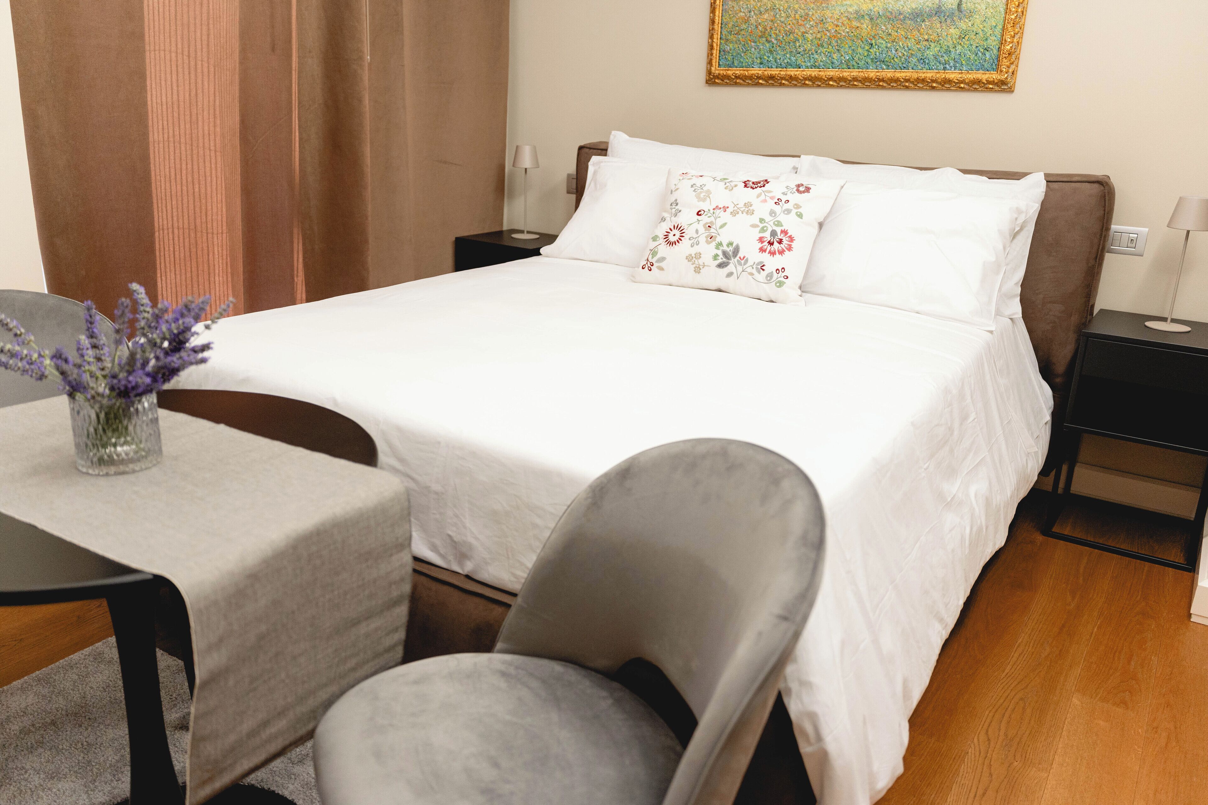 A neatly made bed with white linens and decorative pillows, a small table with a vase of flowers, and a chair.