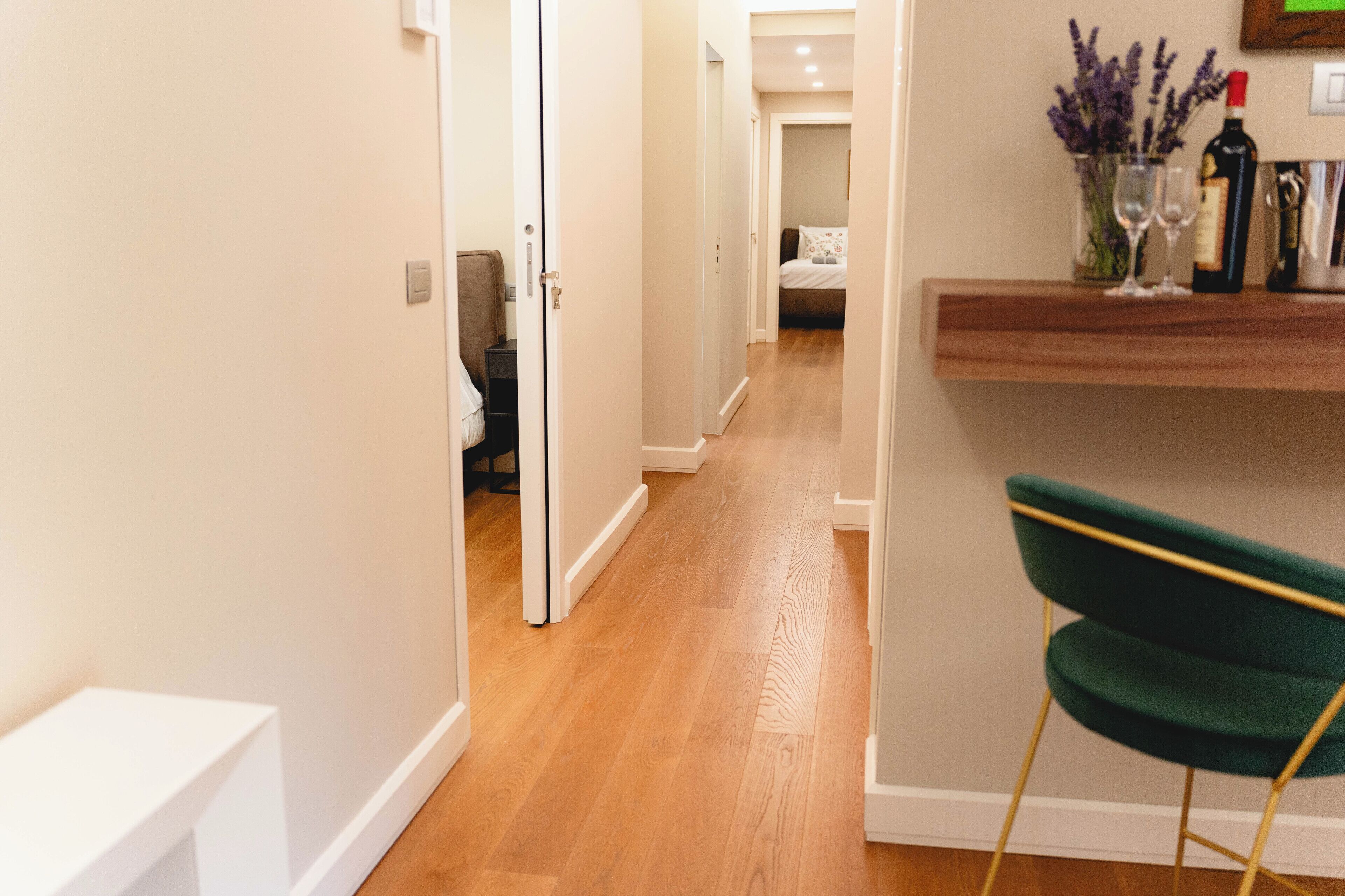 A hallway with wooden flooring, a green chair, and a shelf with a bottle and glasses.