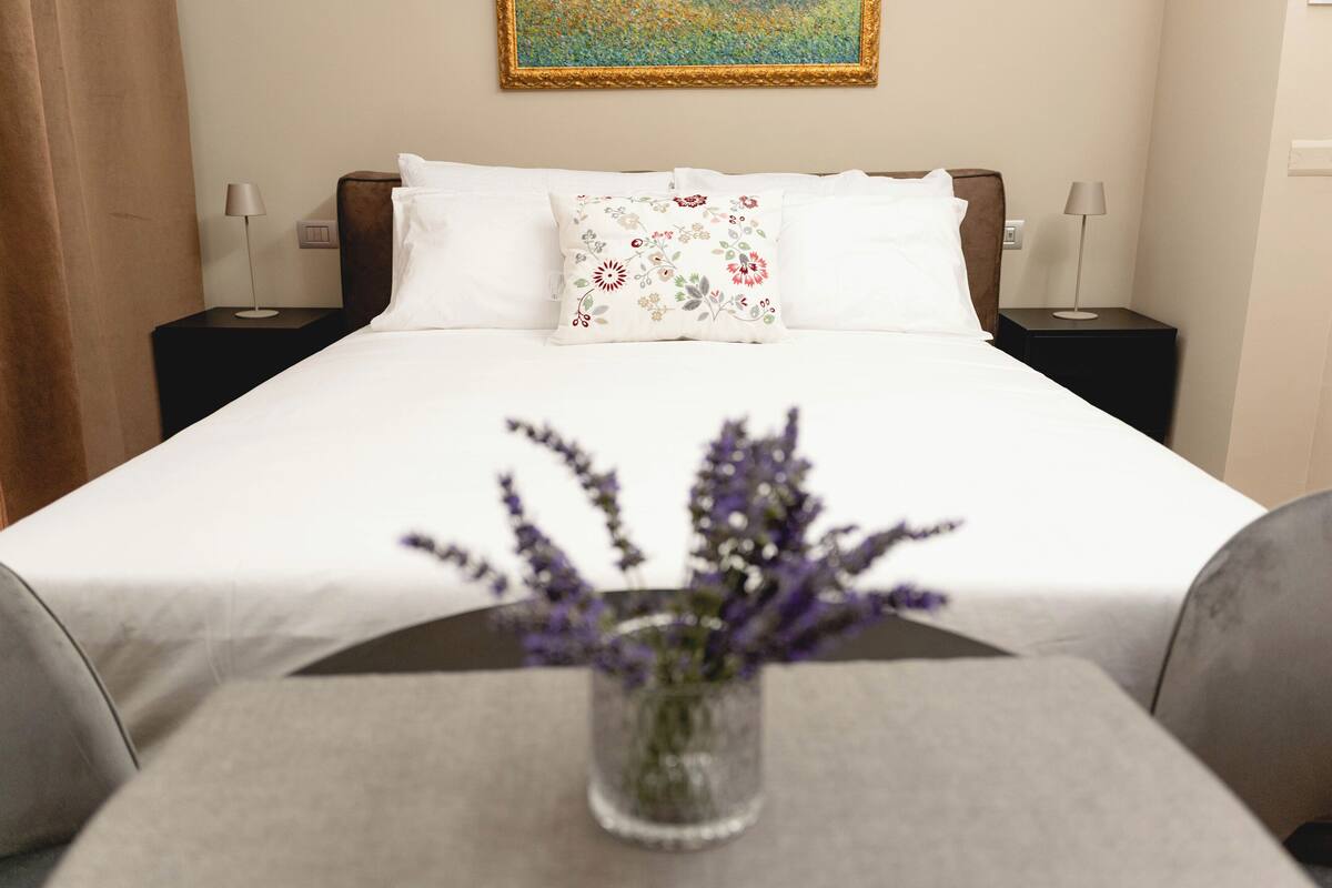 A neatly made bed with white linens and a floral pillow, a small table with a vase of flowers, and two bedside lamps on black nightstands.