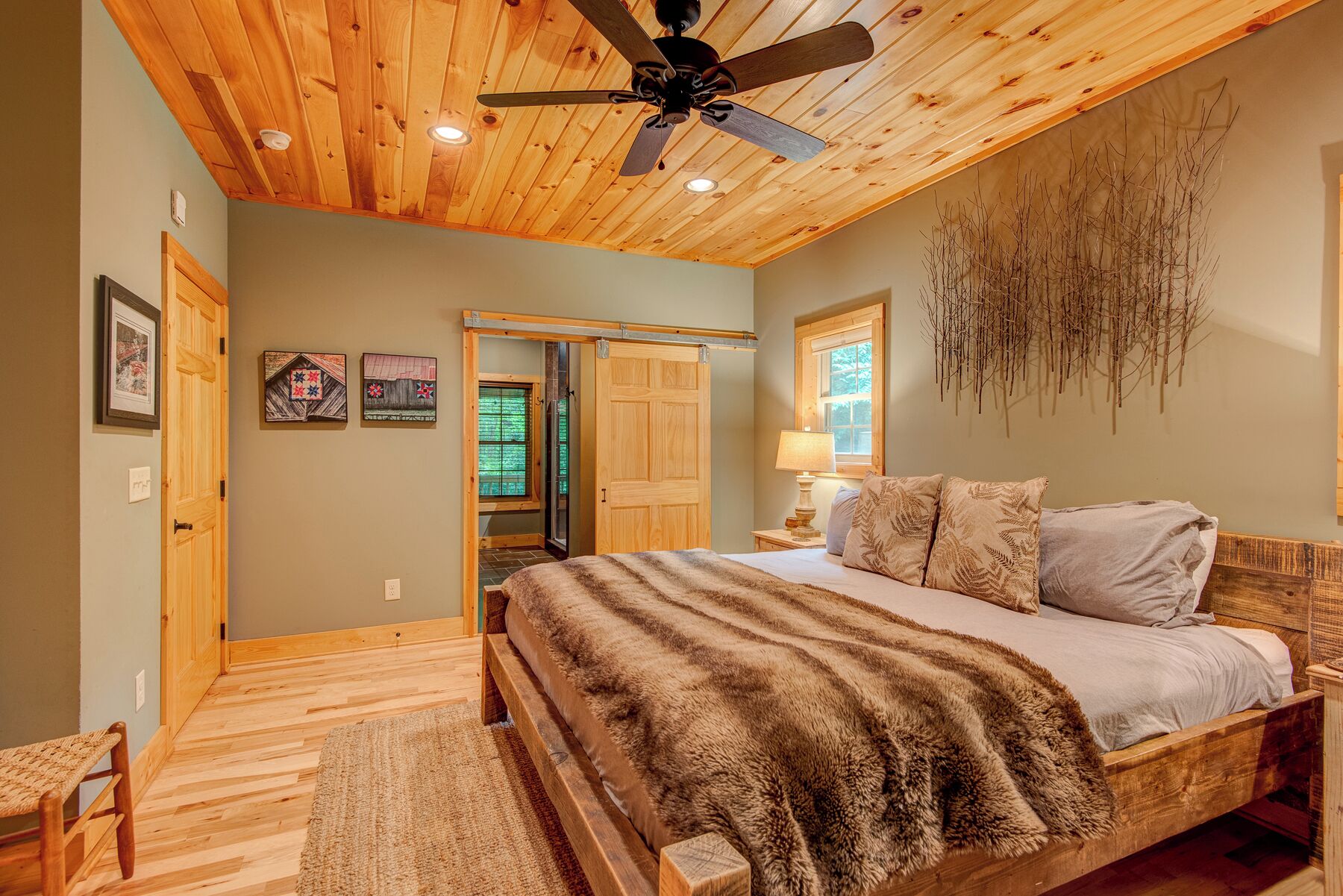A bedroom with a wooden ceiling, a bed with a fur throw, a nightstand with a lamp, and a window with a view of trees.
