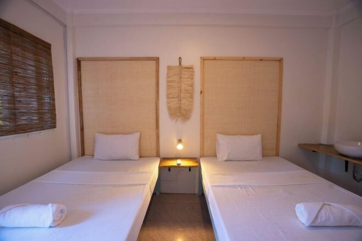 A hotel room with two single beds, a small bedside table with a lamp, a wall-mounted decorative item, and a wooden shelf with a bowl.