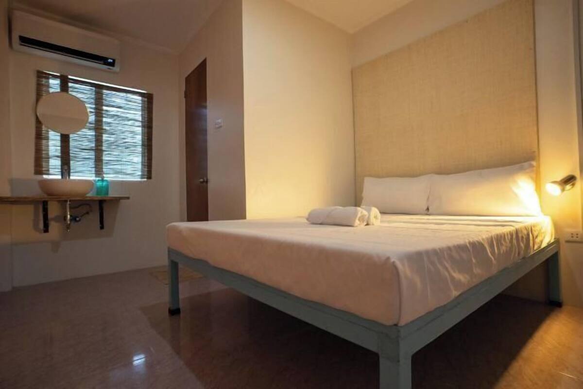 A bedroom with a bed, a bedside table, a window with blinds, and an air conditioning unit.