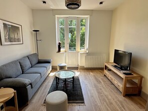 Una sala de estar moderna con un sofá gris, una mesita redonda, un televisor sobre un mueble de madera y un ventanal con vistas a la vegetación.