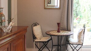 Una habitación con un armario de madera, una silla de metal negro, una mesa redonda con mantel estampado y un espejo en la pared.