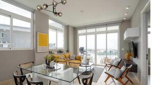 Ein modernes Wohnzimmer mit einem Glastisch, bequemen Sitzgelegenheiten, einem großen Fenster mit Aussicht und einer zeitgemäßen Pendelleuchte.