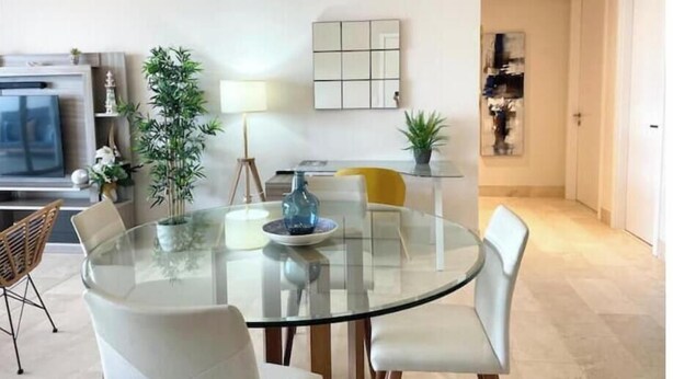 Een moderne woonkamer met een glazen eettafel, witte stoelen, een televisie en decoratieve planten.