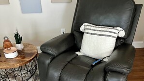 A black leather recliner with a white pillow and a small blue tag, positioned on a patterned rug in a room with a wooden floor and a round coffee table with decorative items. - Cozy sunlit apartment steps from downtown Dover. Parking included (Rollinsford)