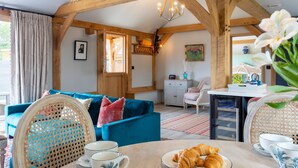 A cozy living room with a blue sofa, wooden beams, a dining table with croissants, and a view of the outside. - The Lodge at Leigh - Luxury Cottages (Sherborne)