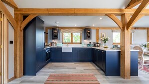 A modern kitchen with dark blue cabinets, wooden beams, and a large window. - The Lodge at Leigh - Luxury Cottages (Sherborne)