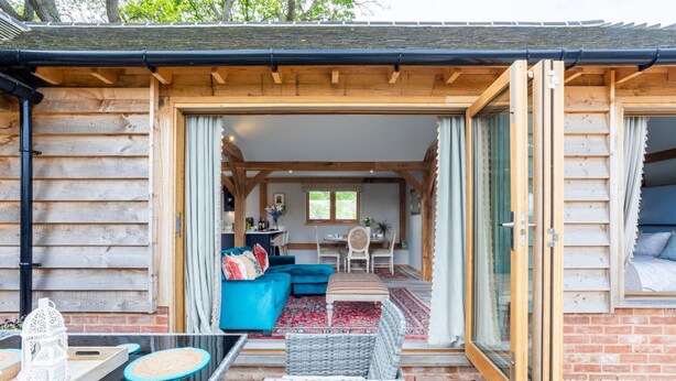 A wooden cabin with an open patio area featuring a sofa, chairs, and a table. - The Lodge at Leigh - Luxury Cottages (Sherborne)