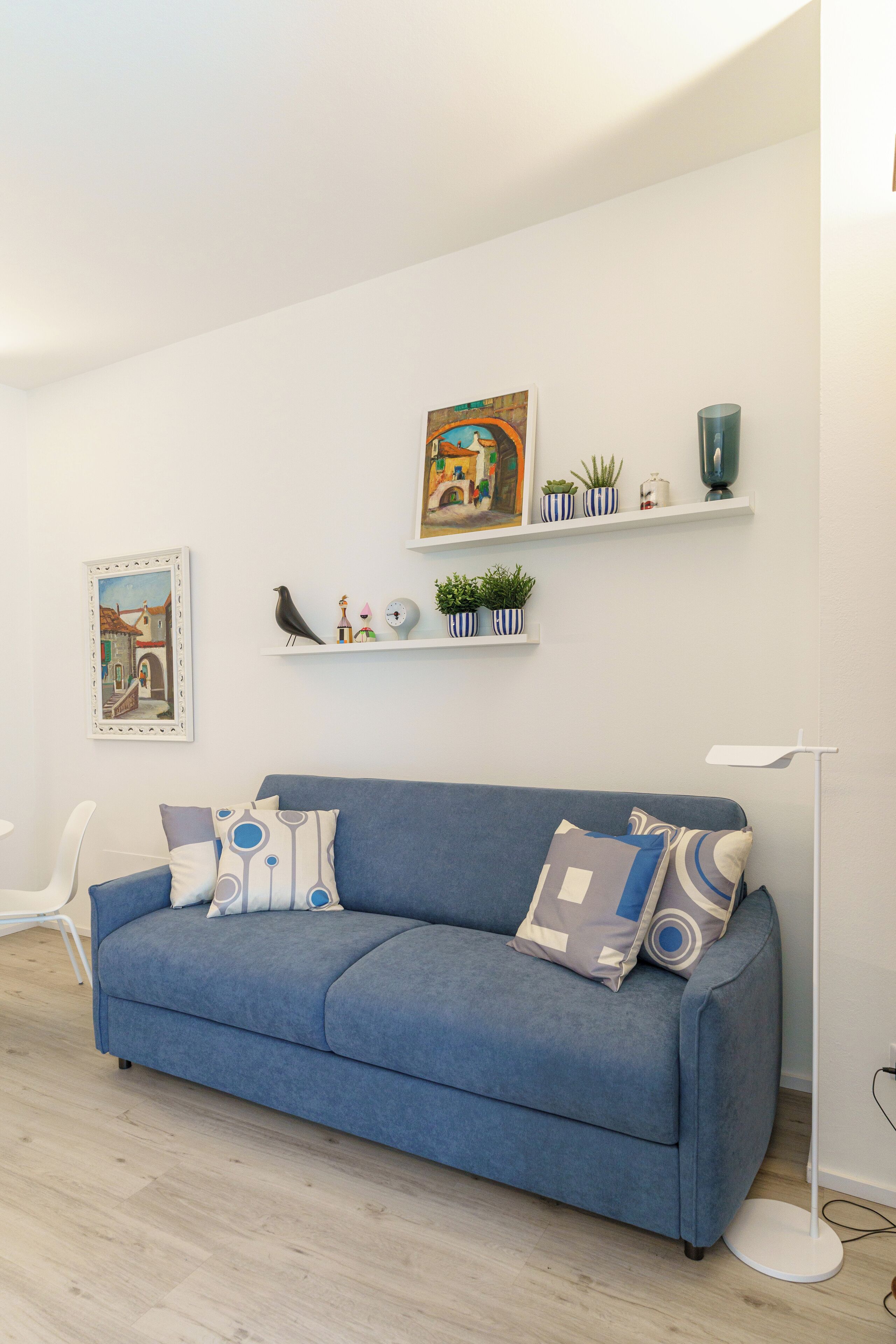 A modern living room with a blue sofa, white walls, and a wooden floor. There are two shelves with decorative items and a framed picture on the wall.