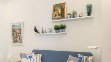 A modern living room with a blue sofa, white walls, and a wooden floor. There are two shelves with decorative items and a framed picture on the wall.
