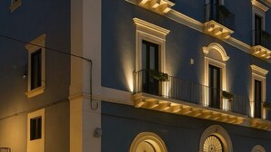 Un immeuble à plusieurs étages, de style architectural classique, doté d’un balcon au deuxième étage et d’un escalier menant à l’entrée.