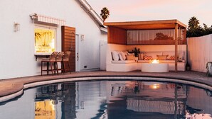 Piscina com deck de madeira, área de estar coberta com almofadas e janela com vista para a piscina.